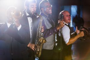A jazz band performing at a nightclub. The four male musicians are standing together, playing a trumpet, saxophone, and tambourine. A mature, African American man is the lead singer, singing into a microphone.